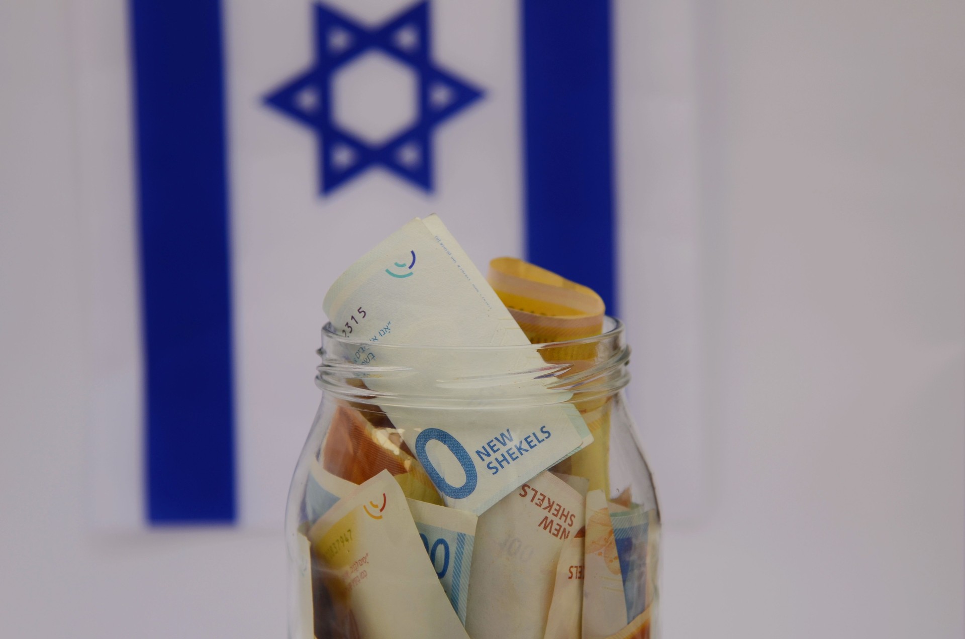 Glass jar with Israeli shekel on the background of the flag of Israel.